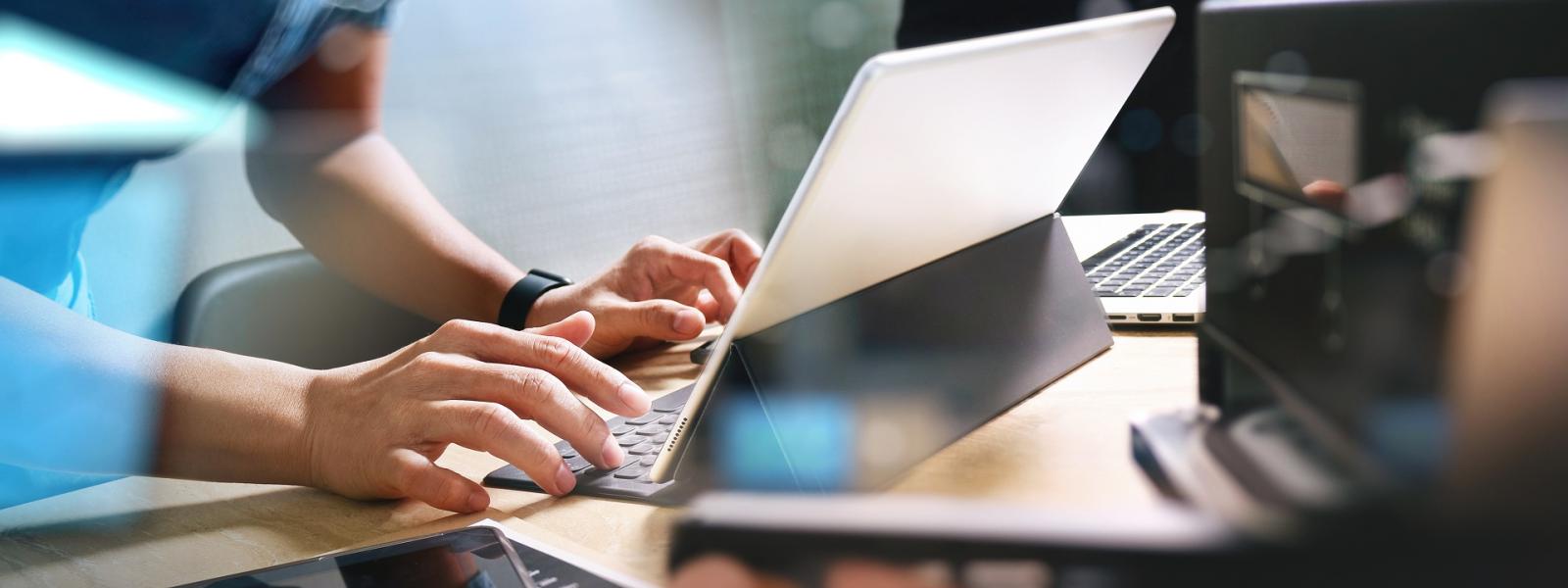 Hands working at computer with person holding cell phone in background